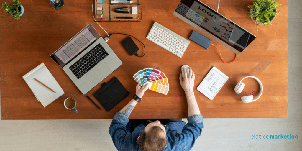 Joven sentado en un escritorio trabajando con un ordenador y colores, buscando una imagen de marca. La imagen tiene vista cenital.